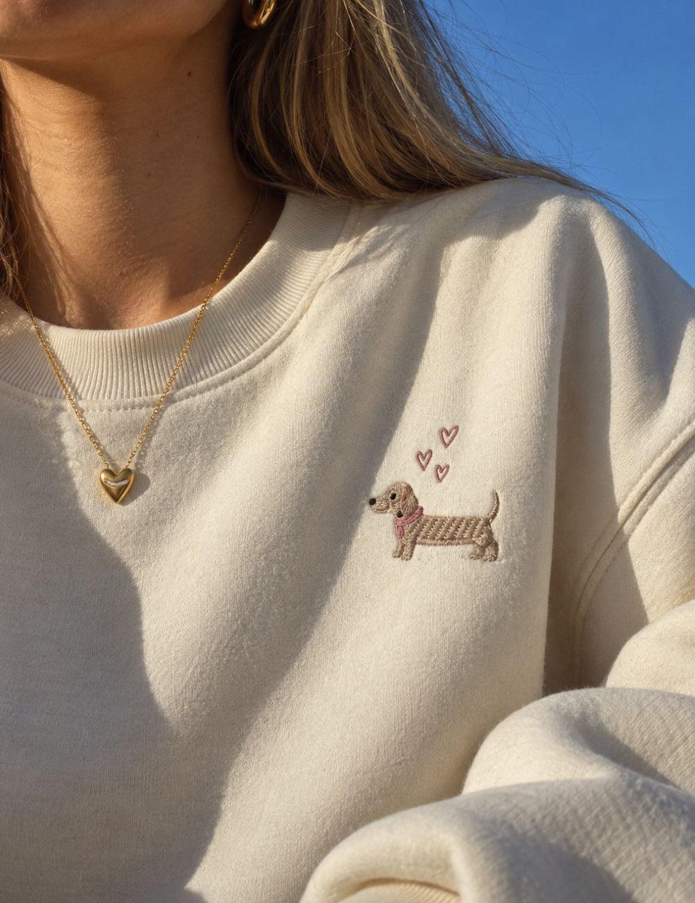 Model wearing a gold heart pendant necklace and classic gold hoop earrings with a cream embroidered sweatshirt.