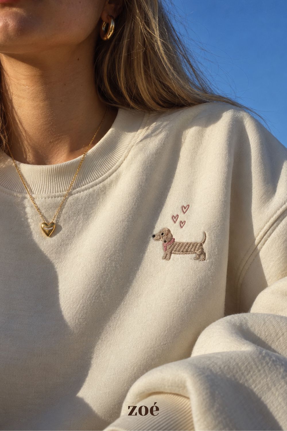 Model wearing a gold heart pendant necklace and classic gold hoop earrings with a cream embroidered sweatshirt.