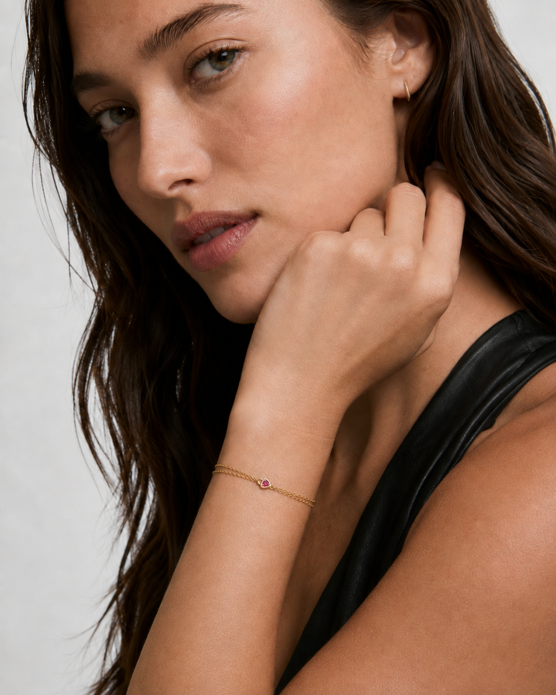 Model wearing a dainty double gold chain bracelet with a pink cubic zirconia heart on the wrist, 18k gold plating.