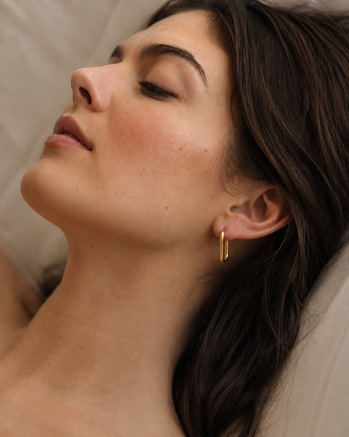 Model wearing an elongated oval gold hoop earring in 18k gold plating, smooth high-polish finish, shown on the ear in soft natural light.