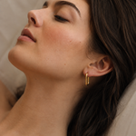 Model wearing an elongated oval gold hoop earring in 18k gold plating, smooth high-polish finish, shown on the ear in soft natural light.