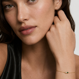 Model wearing a dainty gold chain bracelet with a clear and green cubic zirconia two-stone detail on the wrist, 18k gold plating.