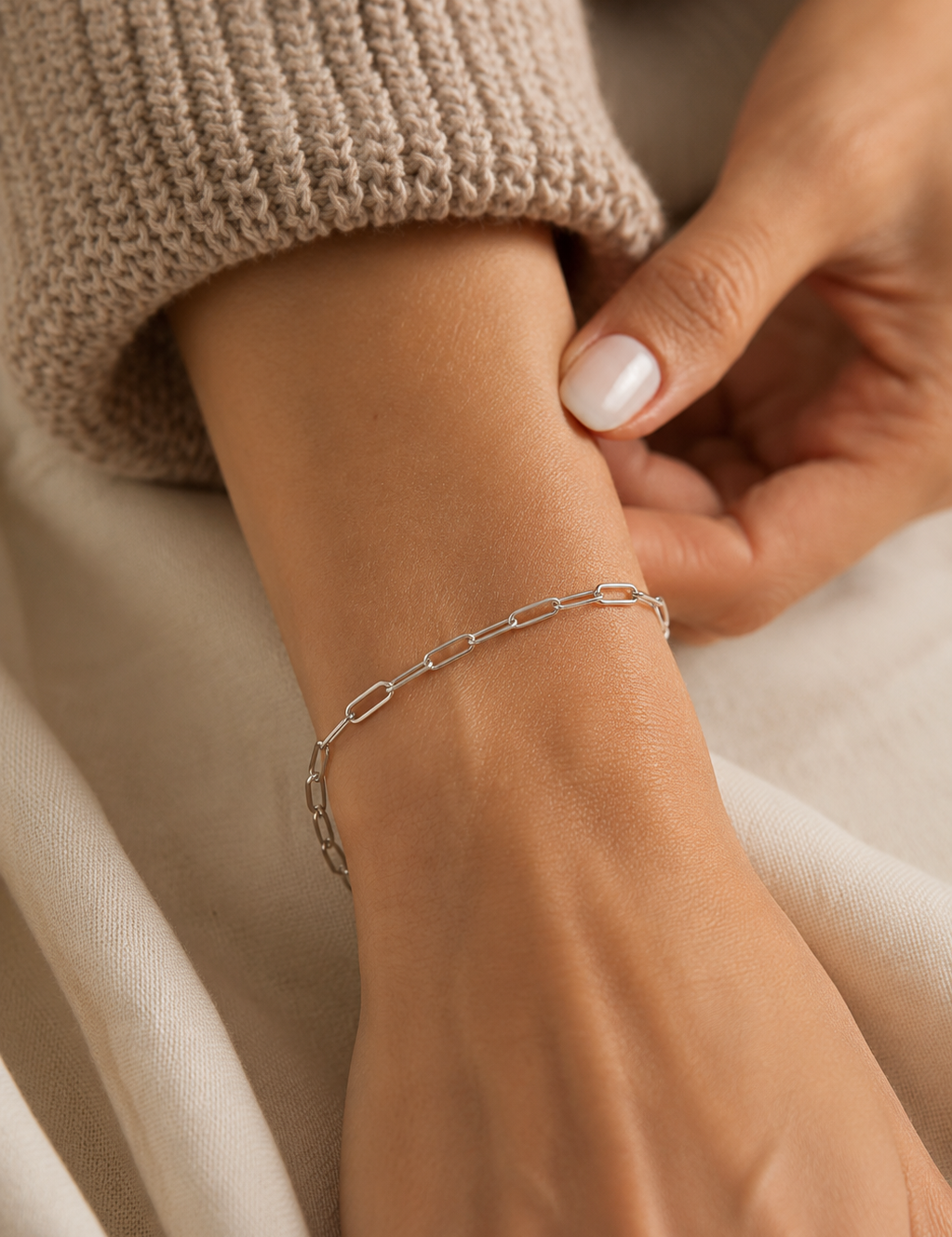 Model wearing a silver-tone paperclip chain bracelet in stainless steel on the wrist, styled with a neutral knit sweater in soft natural light.