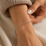 Model wearing a silver-tone paperclip chain bracelet in stainless steel on the wrist, styled with a neutral knit sweater in soft natural light.
